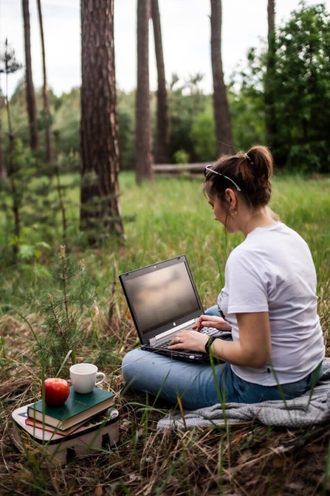 Young woman working remotely in a serene forest setting with laptop, books, and coffee.