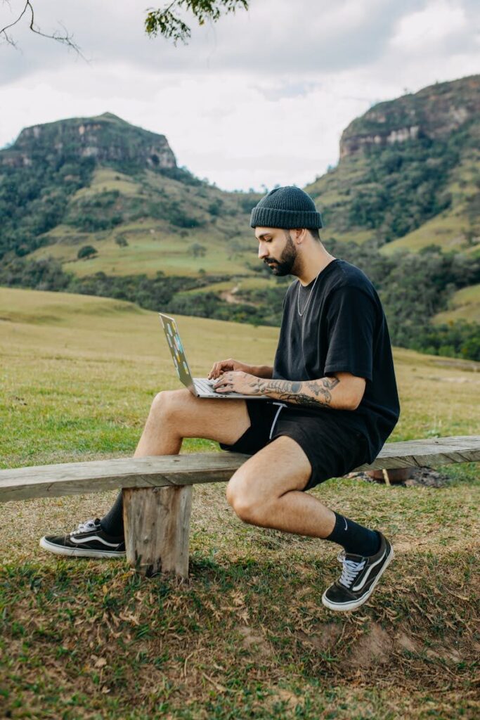 Young man with a beard using a laptop while sitting on a bench in a rural landscape with mountains.